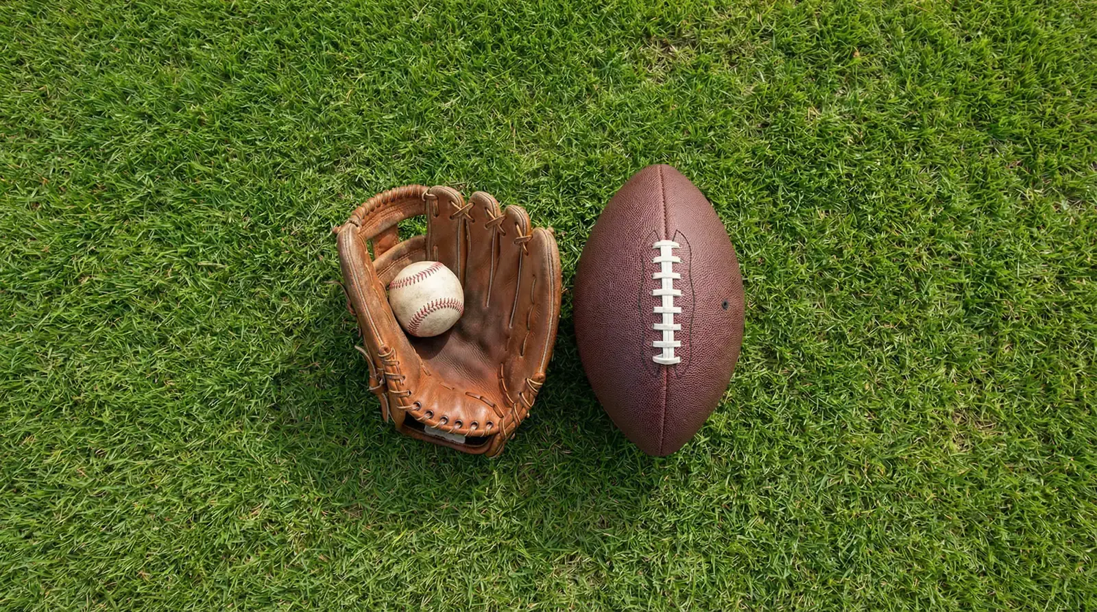 A baseball glove and an American football resting side by side on a grass field under natural daylight